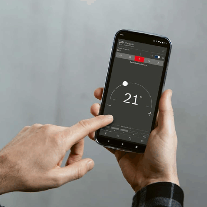 Ein Smartphone wird von zwei Händen gehalten: Die rechte Hand umschließt das Gerät, die linke berührt mit dem Zeigefinger den Bildschirm. Auf dem Display ist die Buderus-Heizungssteuerungs-App zu sehen, die in einem großen, halbkreisförmigen Drehregler die Raumtemperatur von „21 °“ anzeigt. Unter dem Regler befinden sich Plus- und Minus-Buttons zur Anpassung, oben eine Statusleiste mit Menüsymbolen und Informationsanzeige.